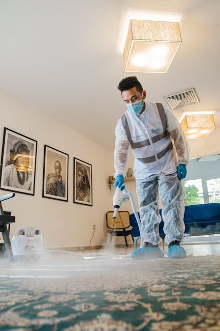 A professional cleaner from Carpet Cleaning Brixton dressed in a white protective suit, blue gloves, and a face mask is performing deep cleaning on a patterned area rug in a modern living room. The cl