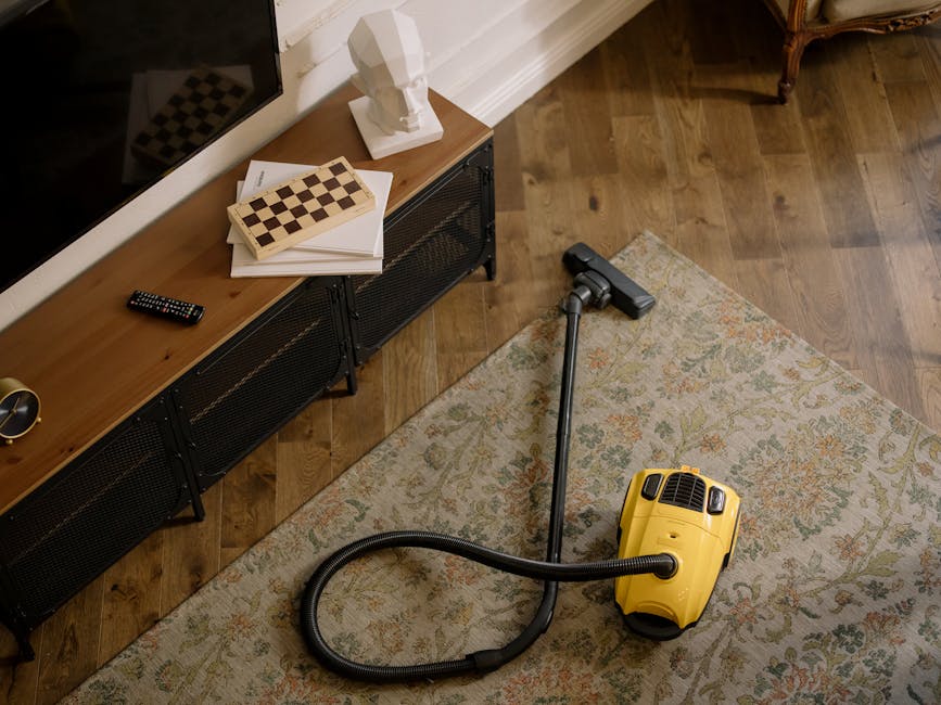 A professional cleaner from Carpet Cleaning Brixton dressed in a white protective suit, blue gloves, and a face mask is performing deep cleaning on a patterned area rug in a modern living room. The cl