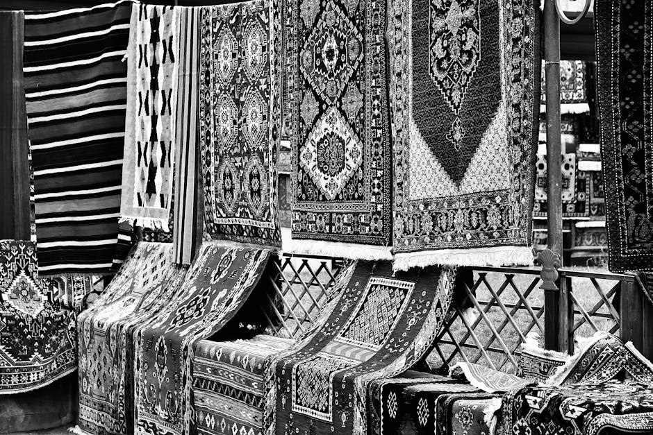 An arrangement of traditional woven rugs and carpets featuring intricate geometric and floral patterns in vibrant reds, blues, and earth tones, displayed on a wooden pallet outdoors. The rugs vary in 